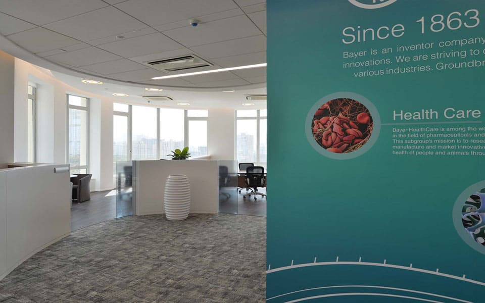 Office reception with curved counter and low glazed screens beside a teal Bayer information banner