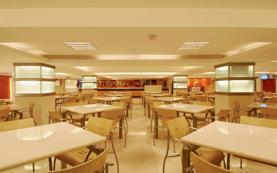 Cafeteria seating rows facing a servery counter, with backlit glass columns and red graphic wall panels