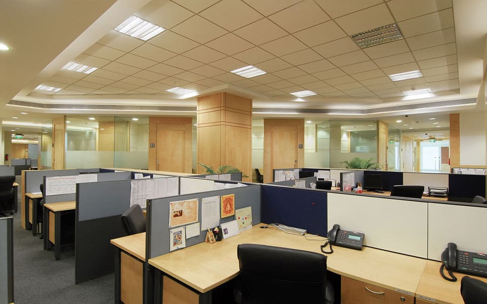 Occupied open-plan office with beech desks, navy tack screens, veneer-clad columns and a louvred fluorescent ceiling