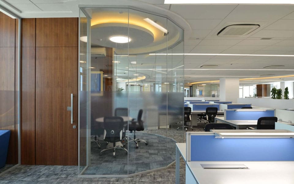 Curved glass meeting room with frosted band and walnut veneer door, beside white bench desks with blue screens