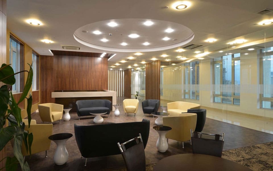 Lounge seating in ochre and charcoal around white pedestal tables, under a circular downlit ceiling disc
