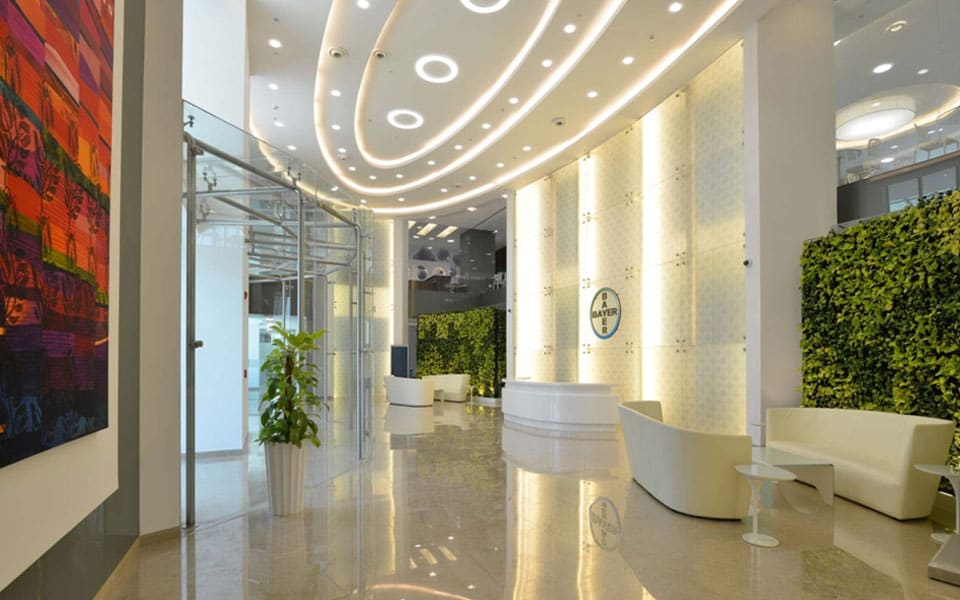 Reception lobby with curved white desk, backlit patterned feature wall carrying the Bayer roundel and green foliage walls
