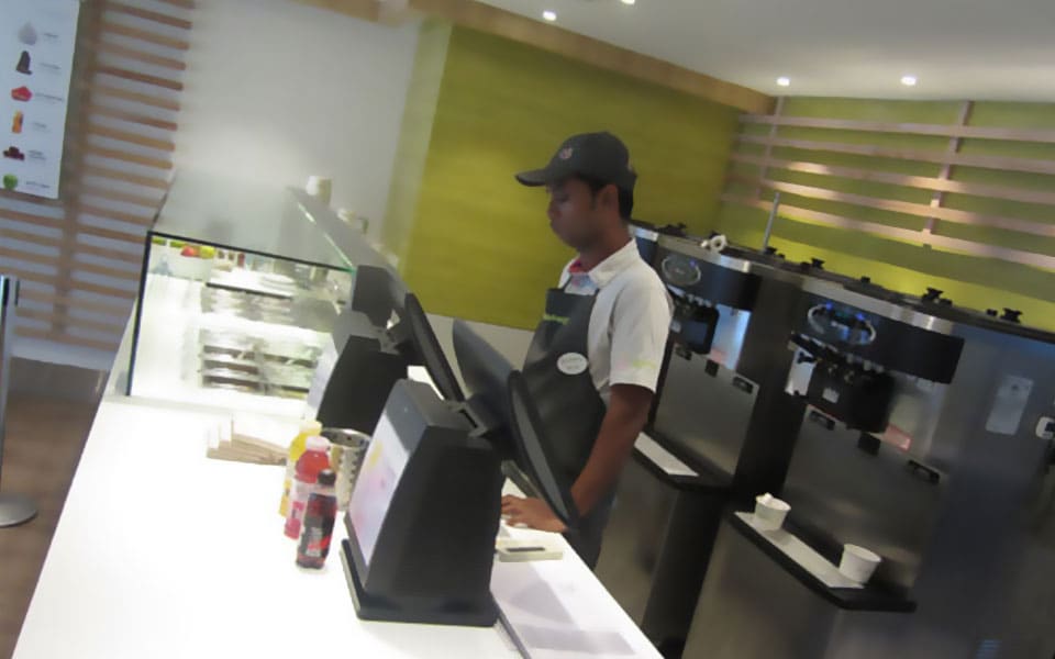 Staff member at the counter beside stainless soft-serve machines and an olive-green feature wall
