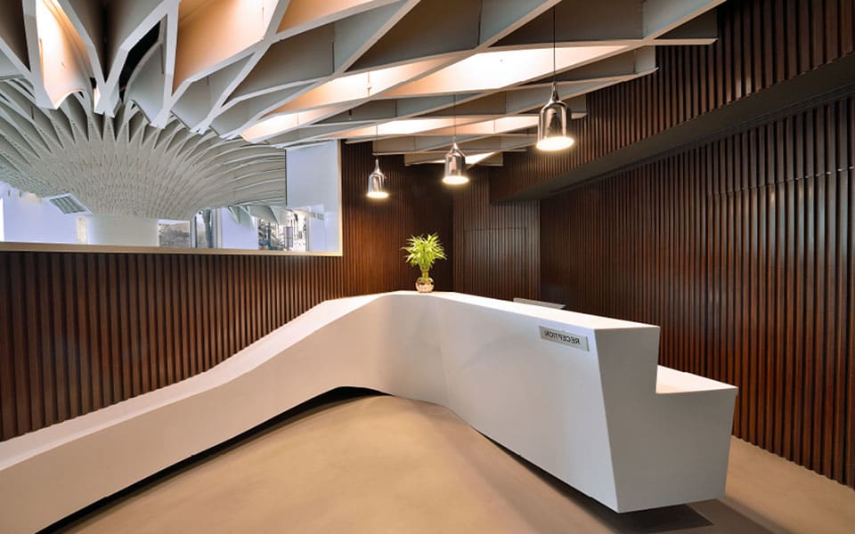 Faceted white solid-surface reception desk against a dark walnut batten wall under three glass pendant lights