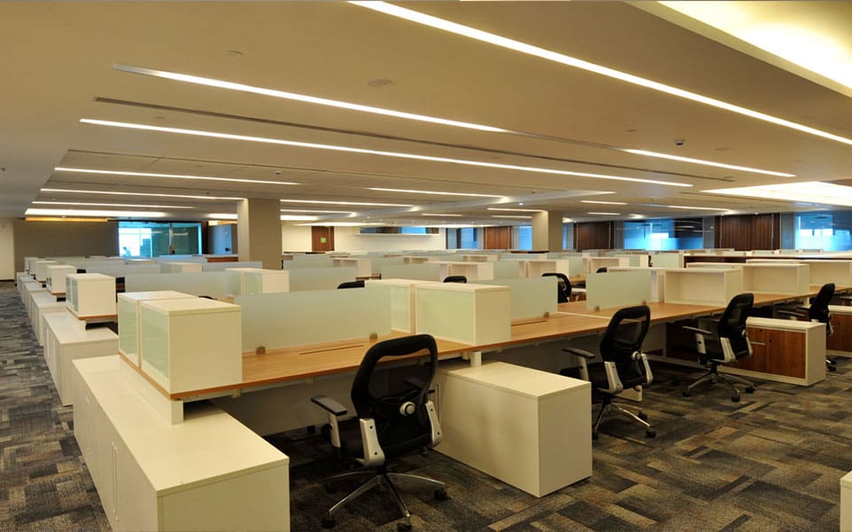 A deep open-plan floor of workstations receding under recessed linear light channels in a ceiling raft