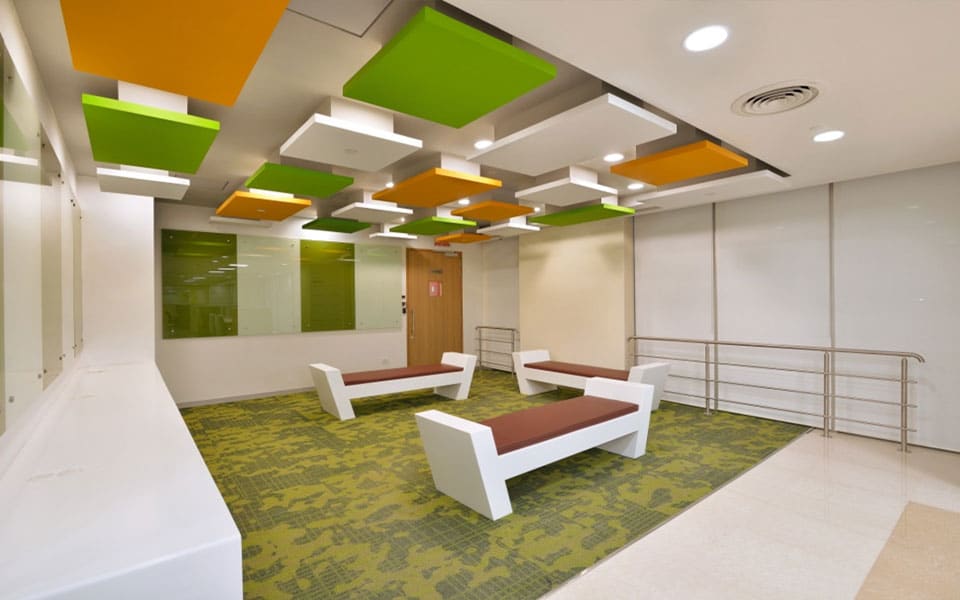 Breakout lounge with white solid-surface benches on green carpet tiles below staggered orange and green acoustic rafts