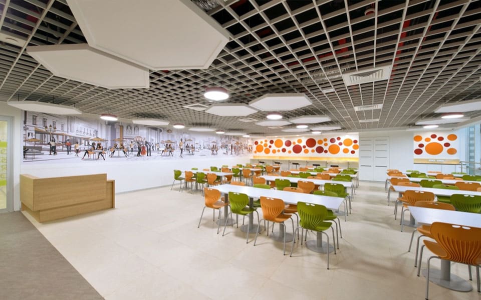 Staff cafeteria with orange and green moulded chairs, a printed street-scene mural and open-cell metal grid ceiling