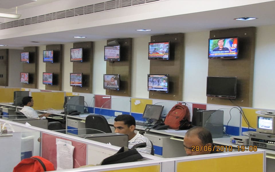 Newsroom workstations below a wall of monitors carrying live Hindi news feeds on veneer panels